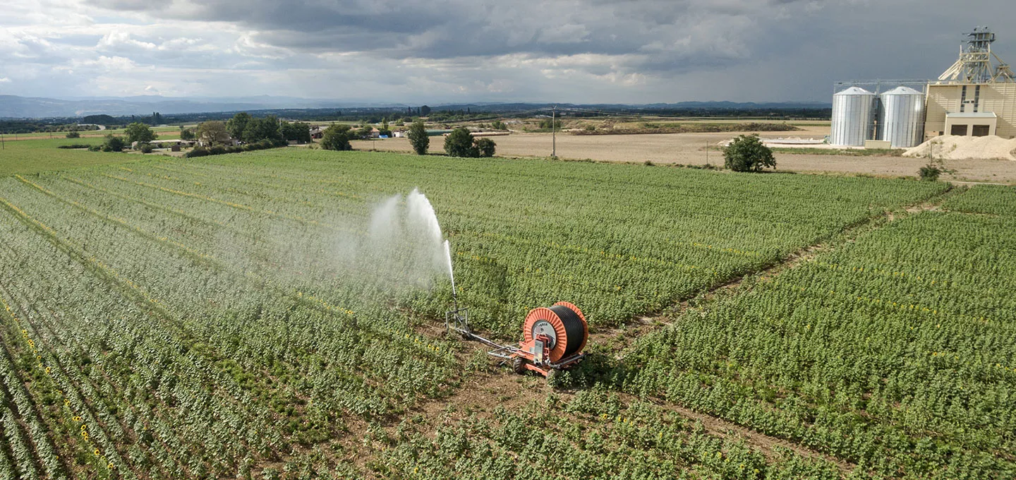 Vue aérienne d’un champ cultivé irrigué par un enrouleur automatique, avec un bâtiment agricole en arrière-plan sous un ciel nuageux.