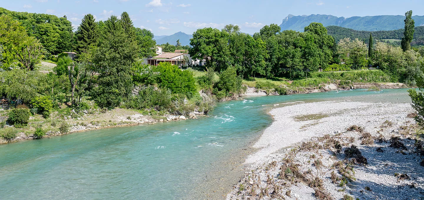 Vue d’une habitation riveraine bordée de végétation, surplombant la rivière Drôme aux eaux turquoise, avec les montagnes des 3 Becs en arrière-plan.