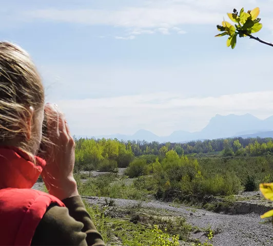 Vue arrière d'une femme en gilet rouge observant la vallée et la rivière Drôme avec des jumelles, face au massif des Trois Becs.