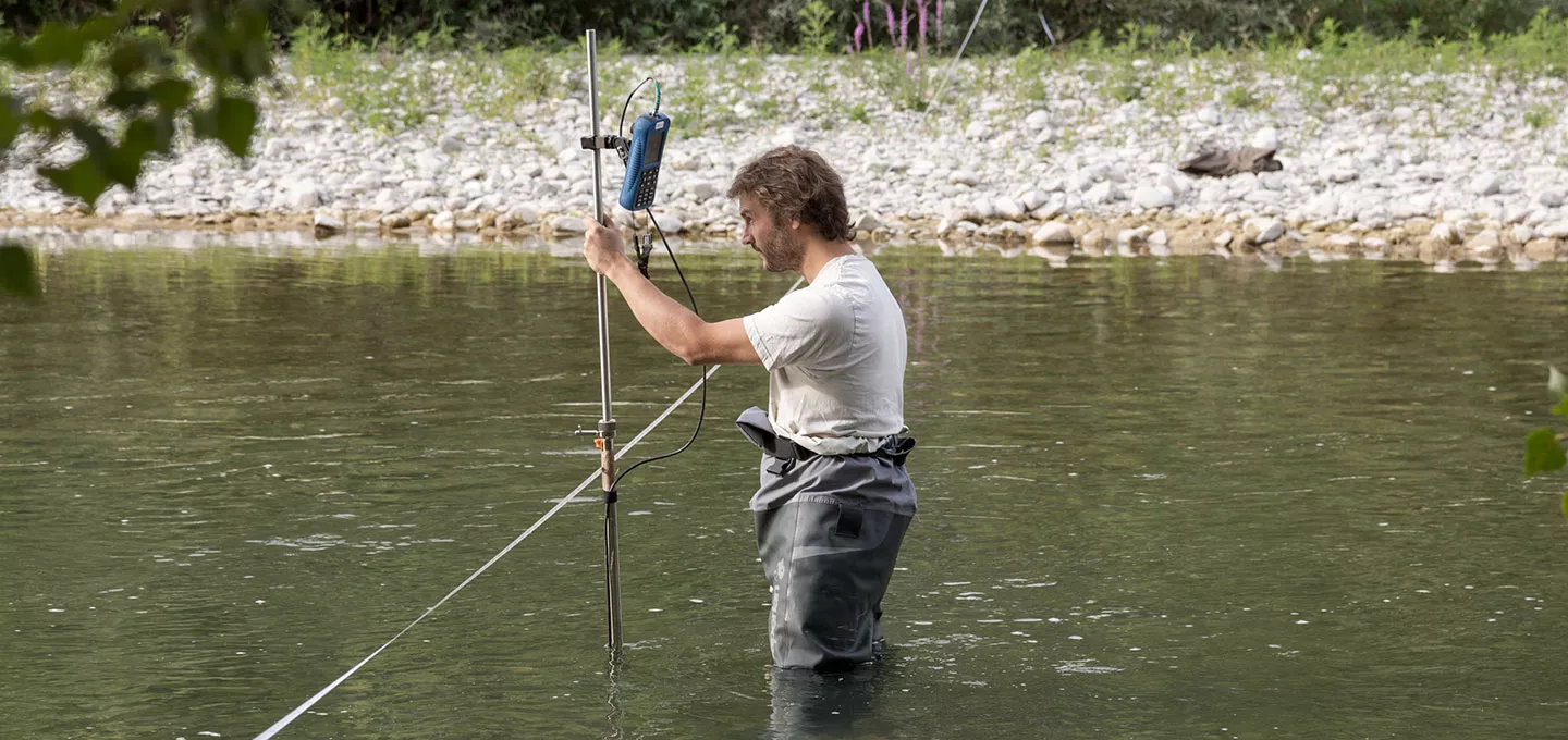 Un technicien équipé de cuissardes réalise des mesures de débit ou de qualité de l’eau dans la rivière Drôme à l’aide d’un appareil de terrain fixé sur une perche.