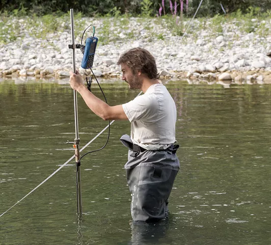 Un technicien en cuissardes installe un capteur dans la rivière Drôme pour mesurer les paramètres hydrologiques, avec un appareil de mesure fixé à une perche en métal.