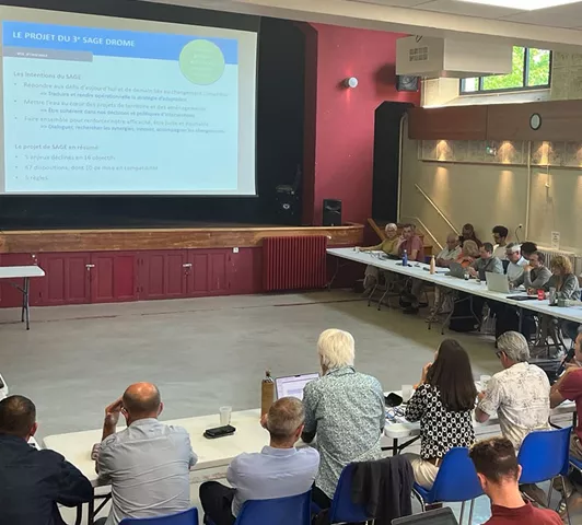 Vue d’une réunion publique avec des participants assis autour de tables en U, assistant à une présentation projetée sur écran au sujet du 3ᵉ SAGE Drôme.