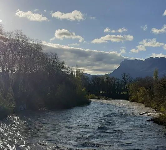 Rivière en crue au coucher du soleil, bordée d’arbres dénudés, avec vue sur les montagnes.