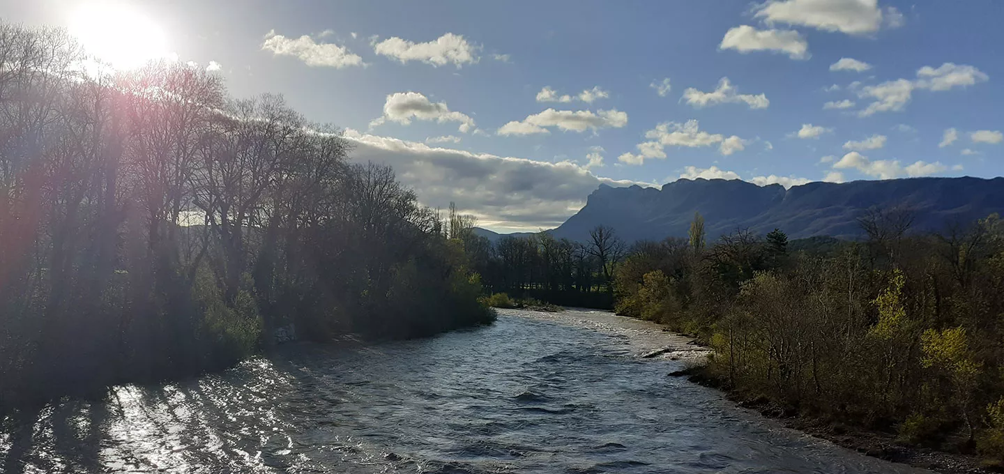Rivière Drôme en crue sous le soleil, avec les arbres en contre-jour et les montagnes en arrière-plan.