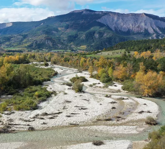 Vue aérienne d’une rivière sinueuse traversant une forêt aux couleurs automnales, avec des montagnes en arrière-plan.