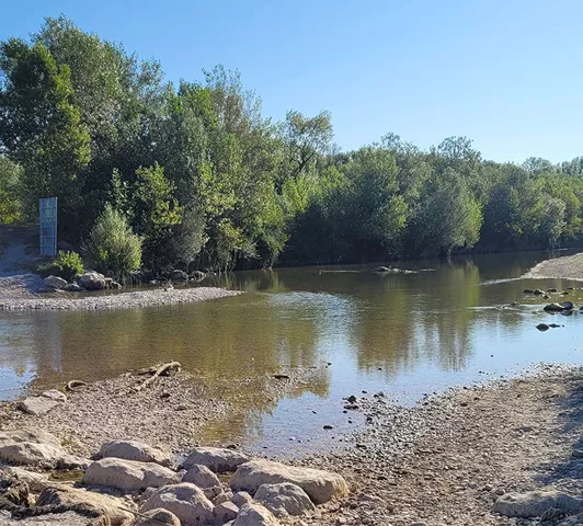 Vue d’un bras peu profond de la rivière Drôme, presque à sec, avec galets, végétation dense en arrière-plan, et panneau de signalisation visible à gauche.