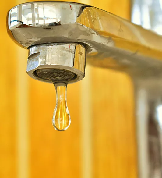 Gros plan sur un robinet métallique d’où s’échappe une goutte d’eau, sur fond jaune flouté.